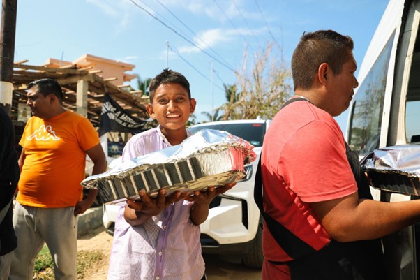 Smiling boy holds a large foil food tray while standing near a man distributing meals from a vehicle.