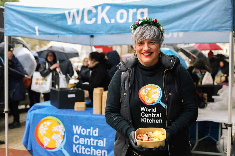 Smiling woman in World Central Kitchen gear and festive headband holds a meal in front of a blue WCK tent.