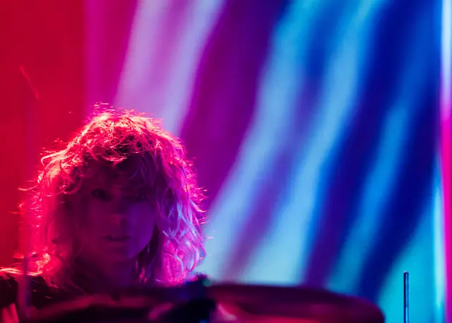 Close-up photo of Stephanie Bailey on drums starring straight ahead in front of strong red and blue light