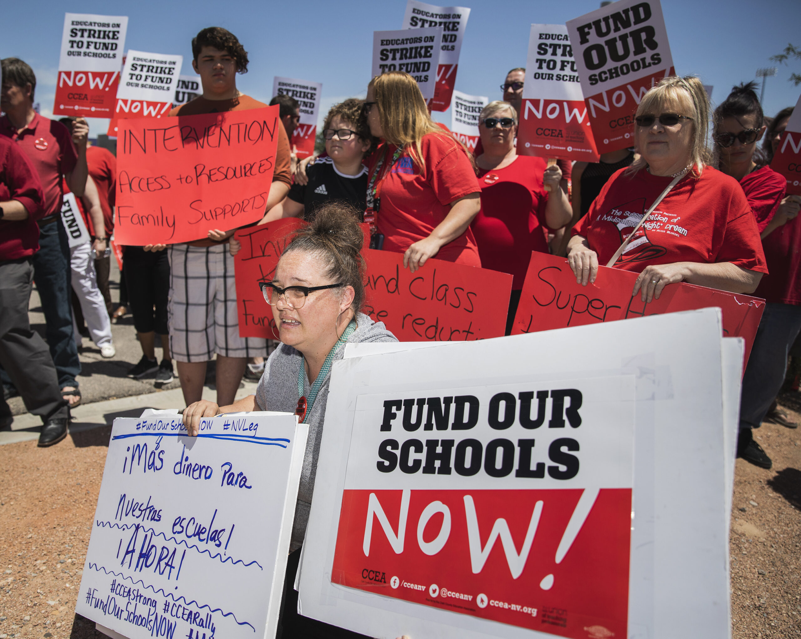 Two teachers with signs kneeling in front a crowd during a rally