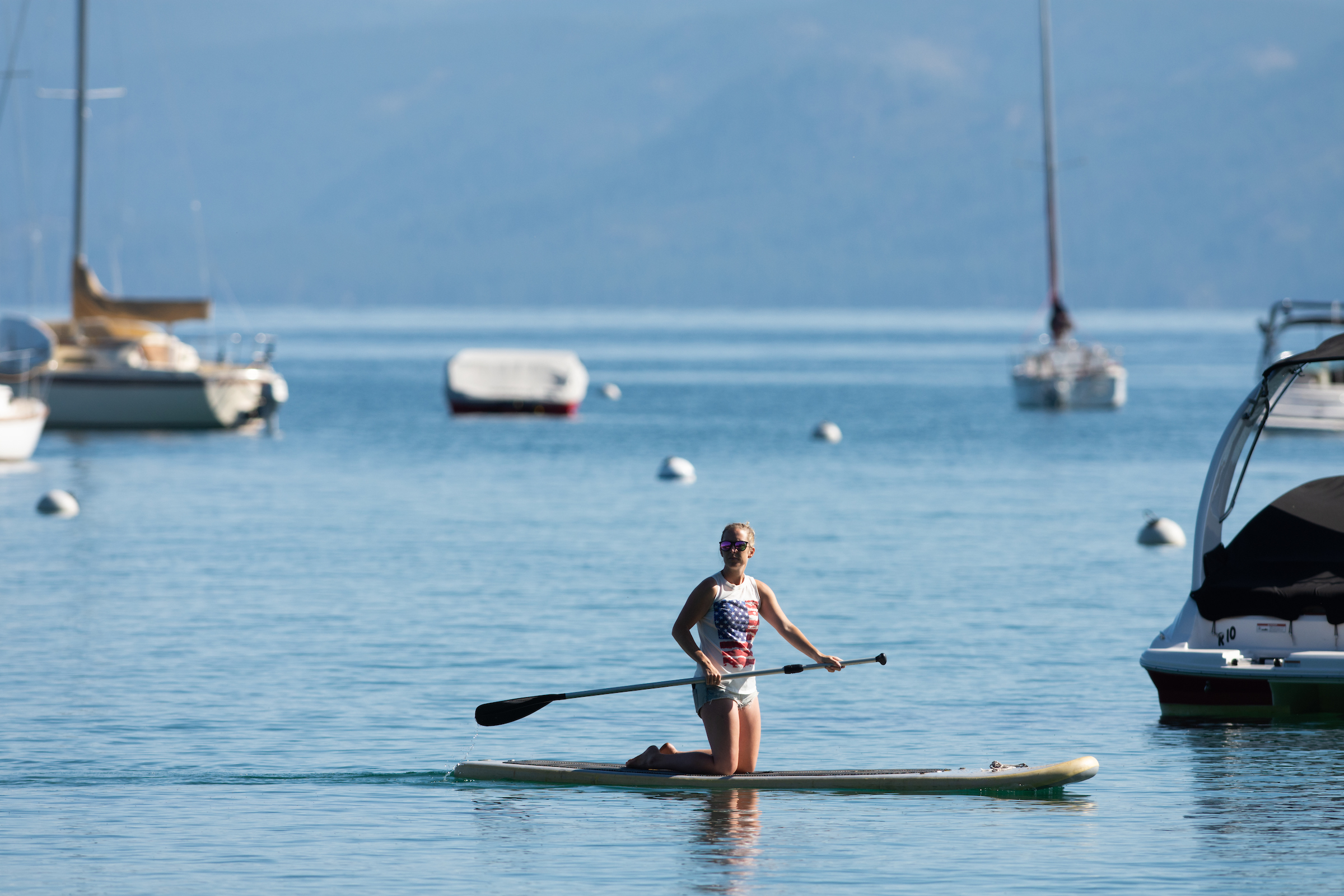 A kneeling paddle boarder in Lake Tahoe with a sailboat in the background