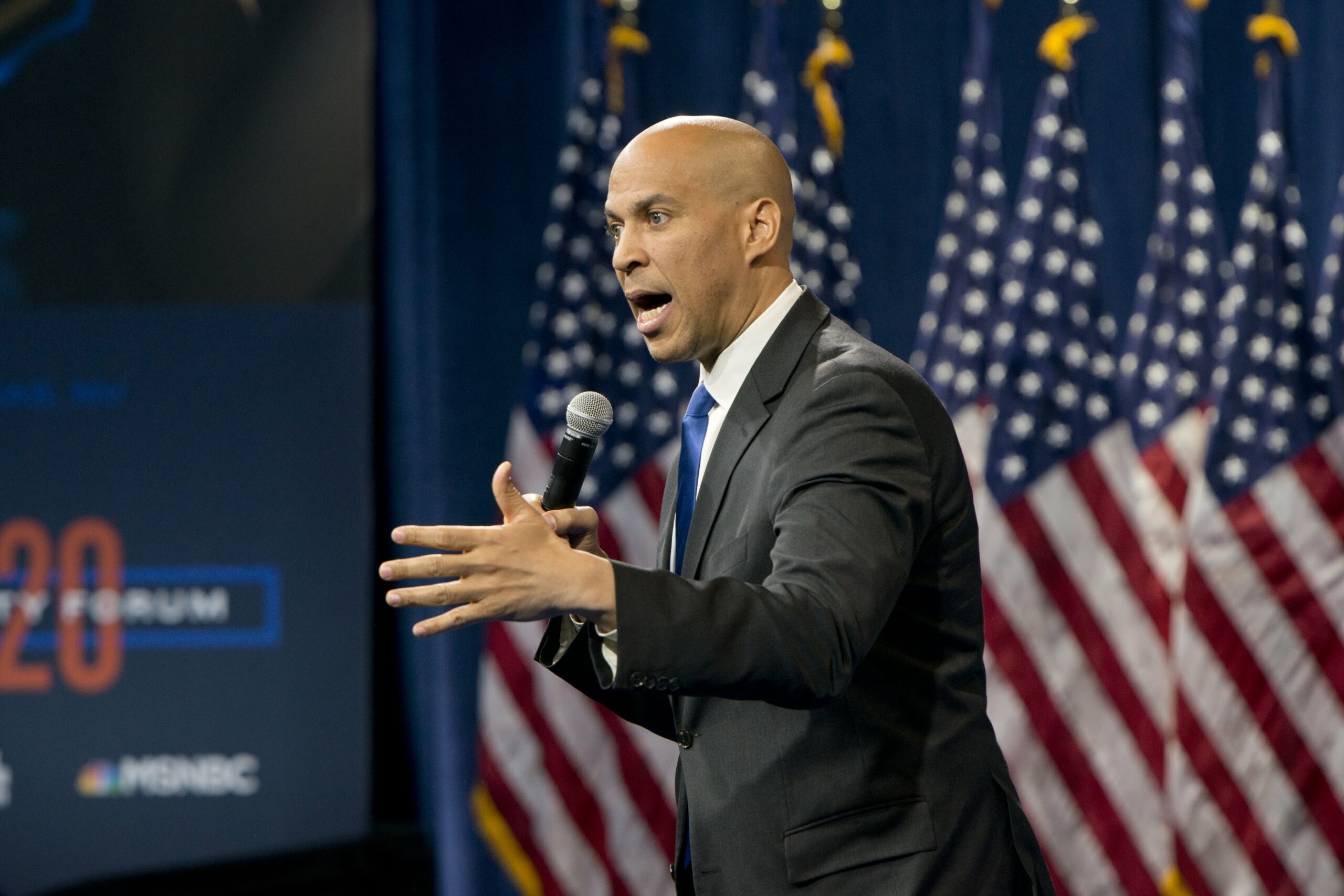 Cory Booker on stage in front of US Flags holding a microphone