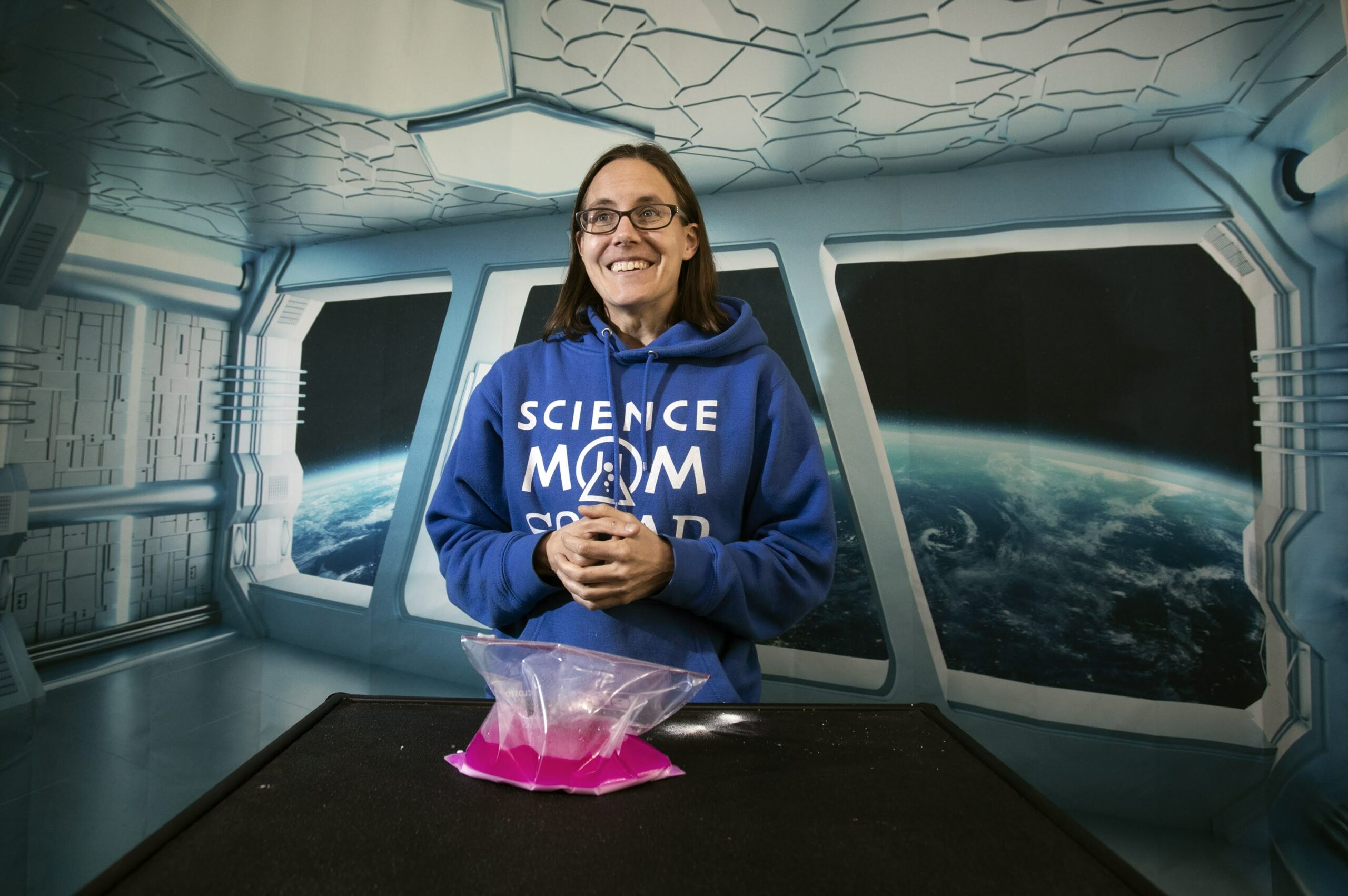 Jenny Ballif, in a blue science mom squad sweatshirt, standing in a space themed room