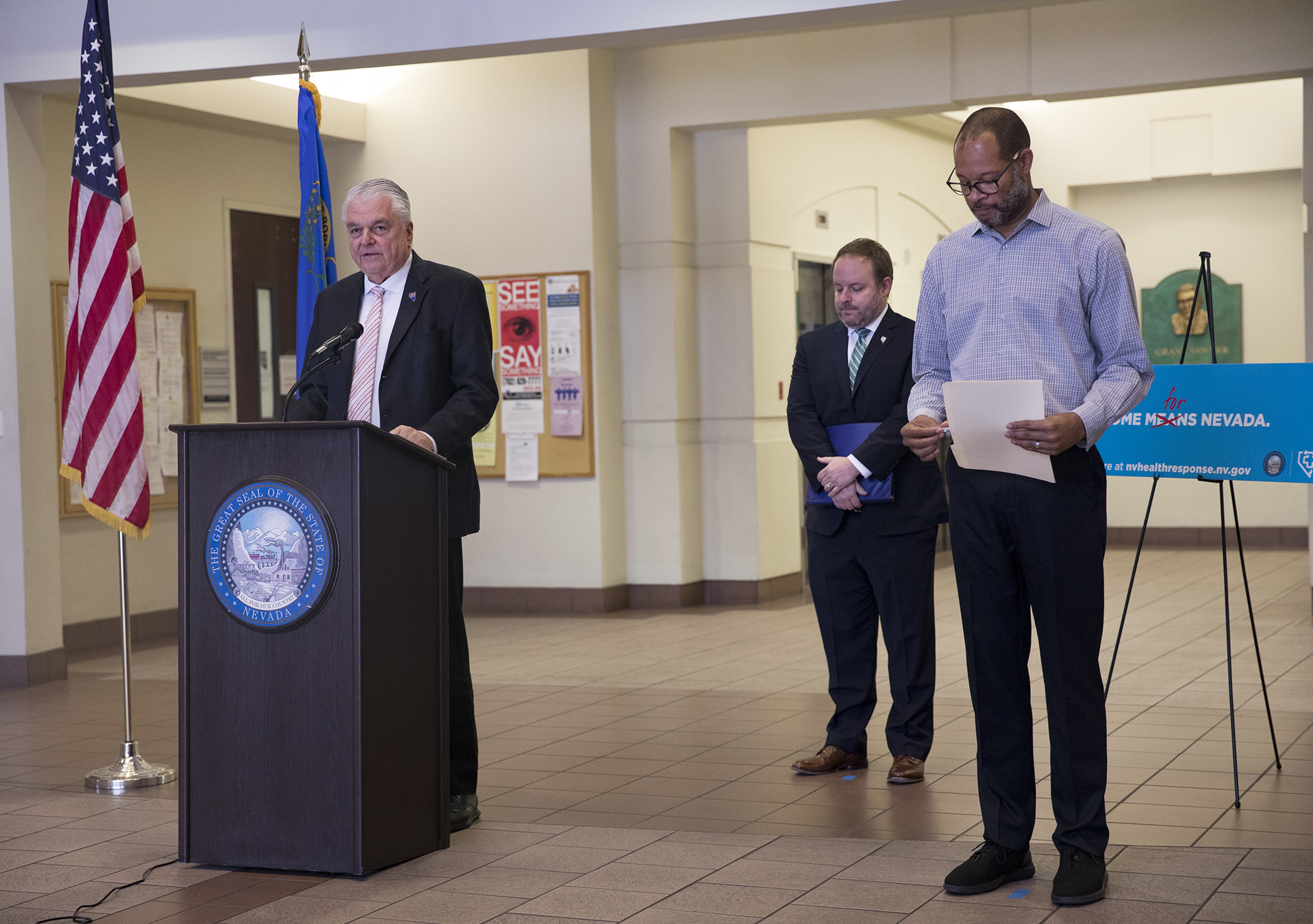 Gov. Steve Sisolak stands behind a podium at a press conference, alongside Treasurer Zach Conine and Attorney General Aaron Ford.