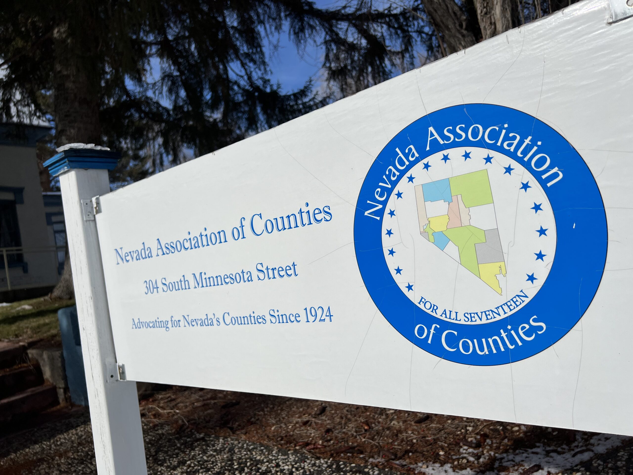 A white rectangular sign outdoors with blue lettering that reads “Nevada Association of Counties 304 South Minnesota Street Advocating For Nevada’s Counties Since 1924.” With a map of Nevada with outlines of the counties inside a blue circle that reads, “Nevada Association of Counties For All 17,” with blue stars.