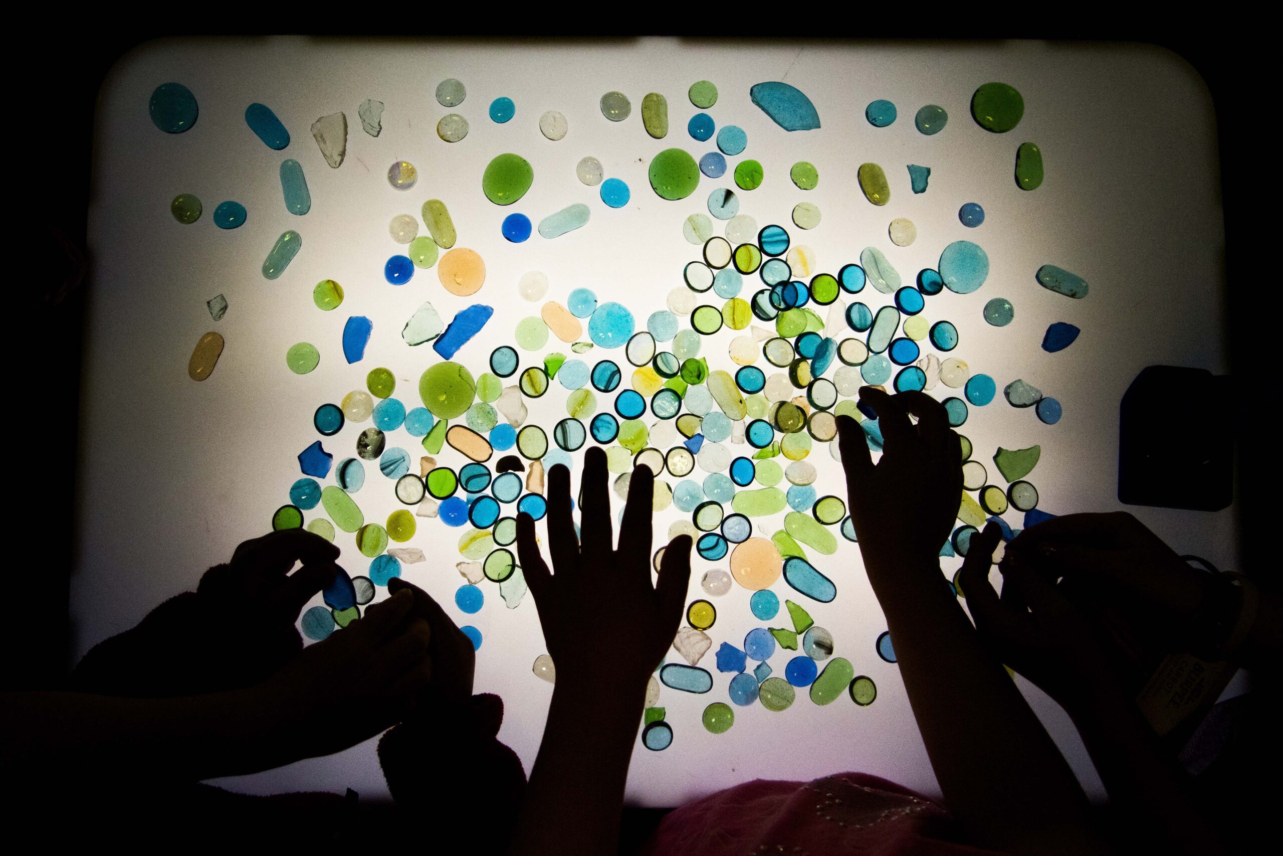 Silhouettes of hands as they move around colored rocks that are lit from underneath.