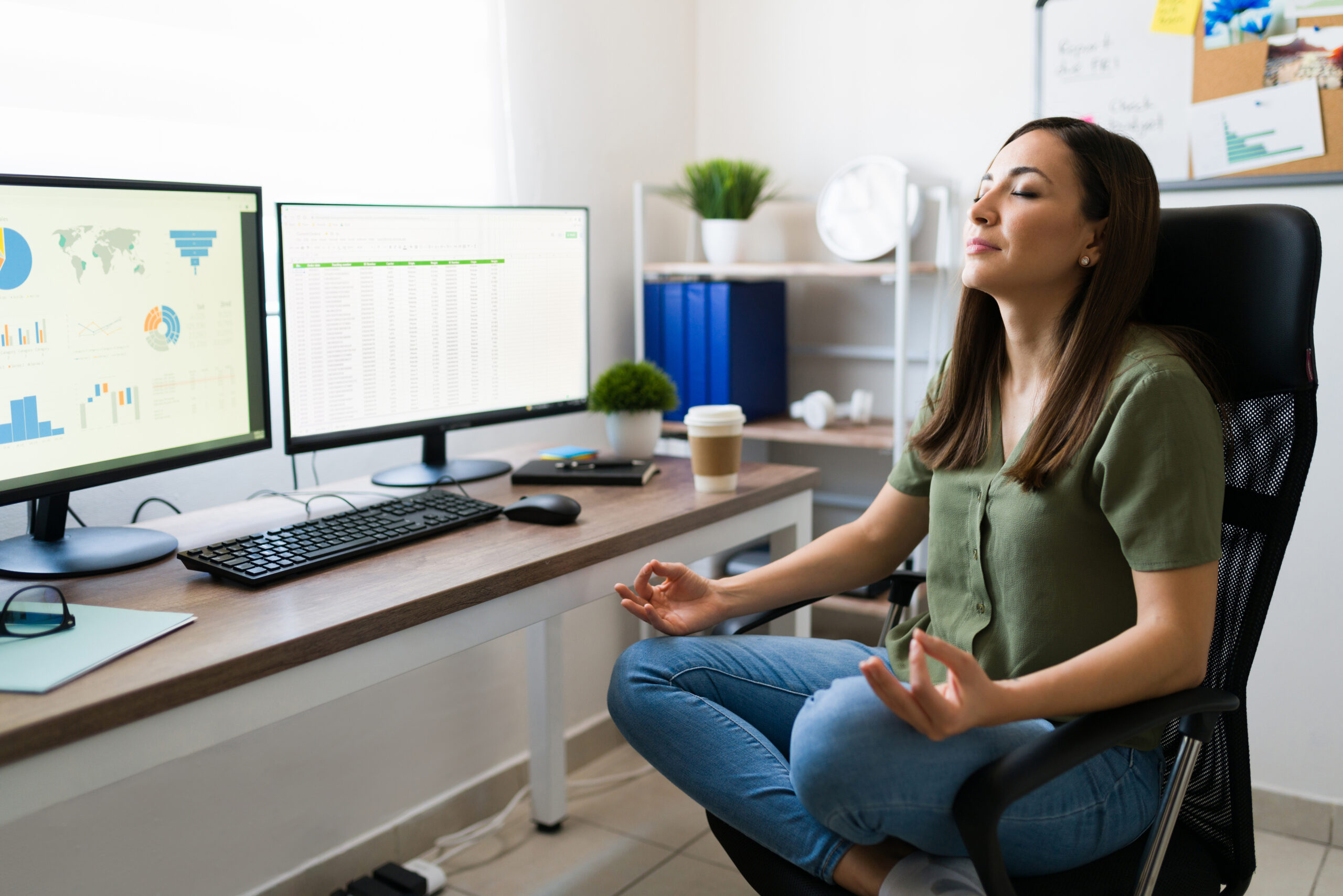 A woman meditating in her office chair in front of her computers.