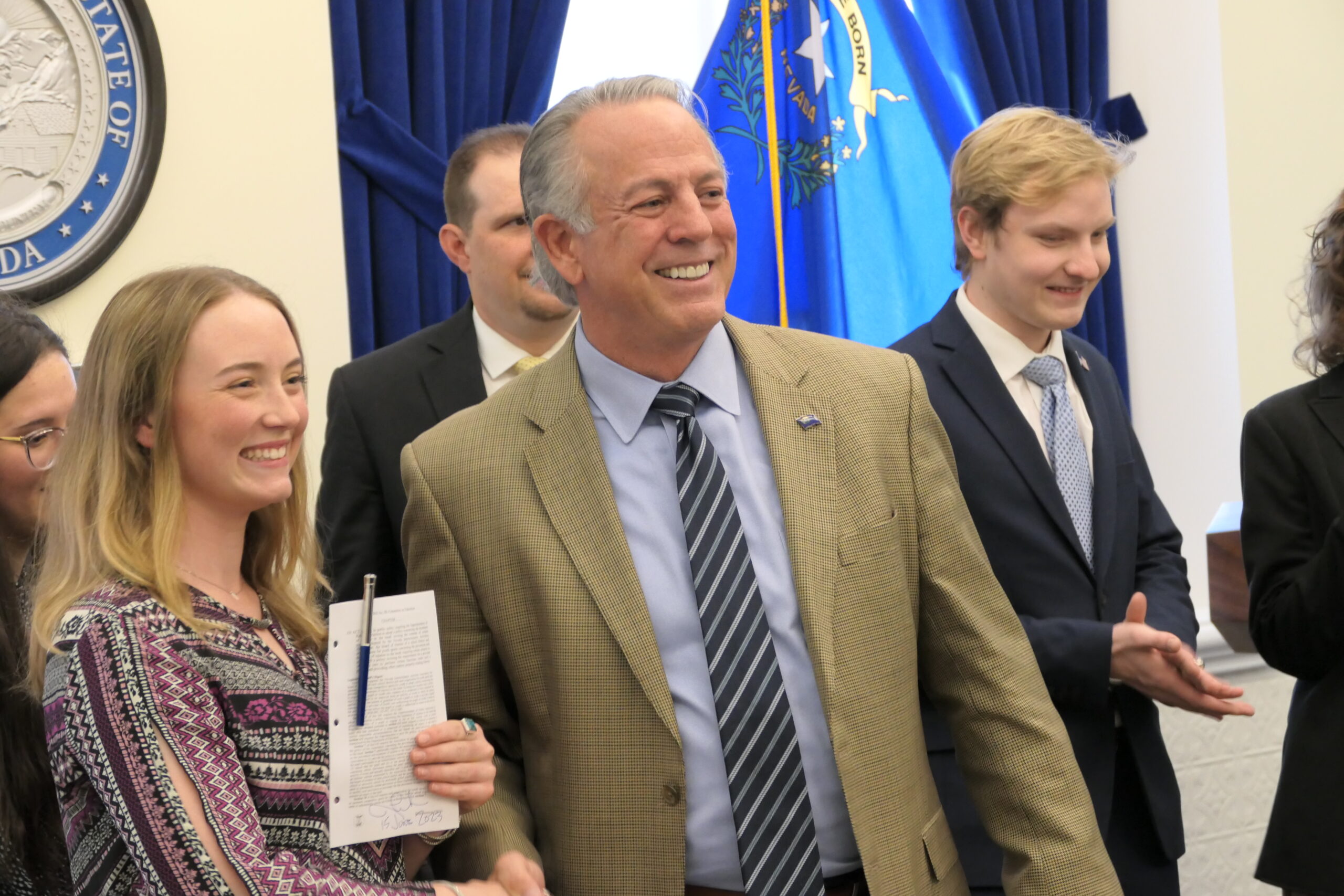 Stella Thornton (left) stands next to Gov. Joe Lombardo (middle) and youth legislator Dillon Moss (back right) at the ceremonial bill signing of SB80 holding a signed copy of SB80 — the bill that Thornton sponsored that updates current law to incorporate procedures for students returning to school after a traumatic brain injury — held in the governor's office in Carson City on Thursday, June 15, 2023. (Carly Sauvageau/The Nevada Independent).