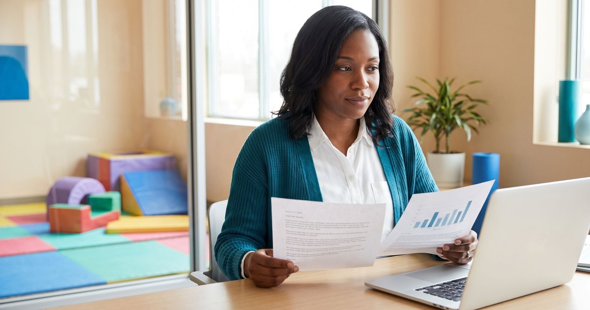 A behavior analyst in a teal cardigan compares two printed job offer letters at her desk in a bright ABA clinic office, with a laptop open and colorful soft play mats visible through the glass wall behind her.