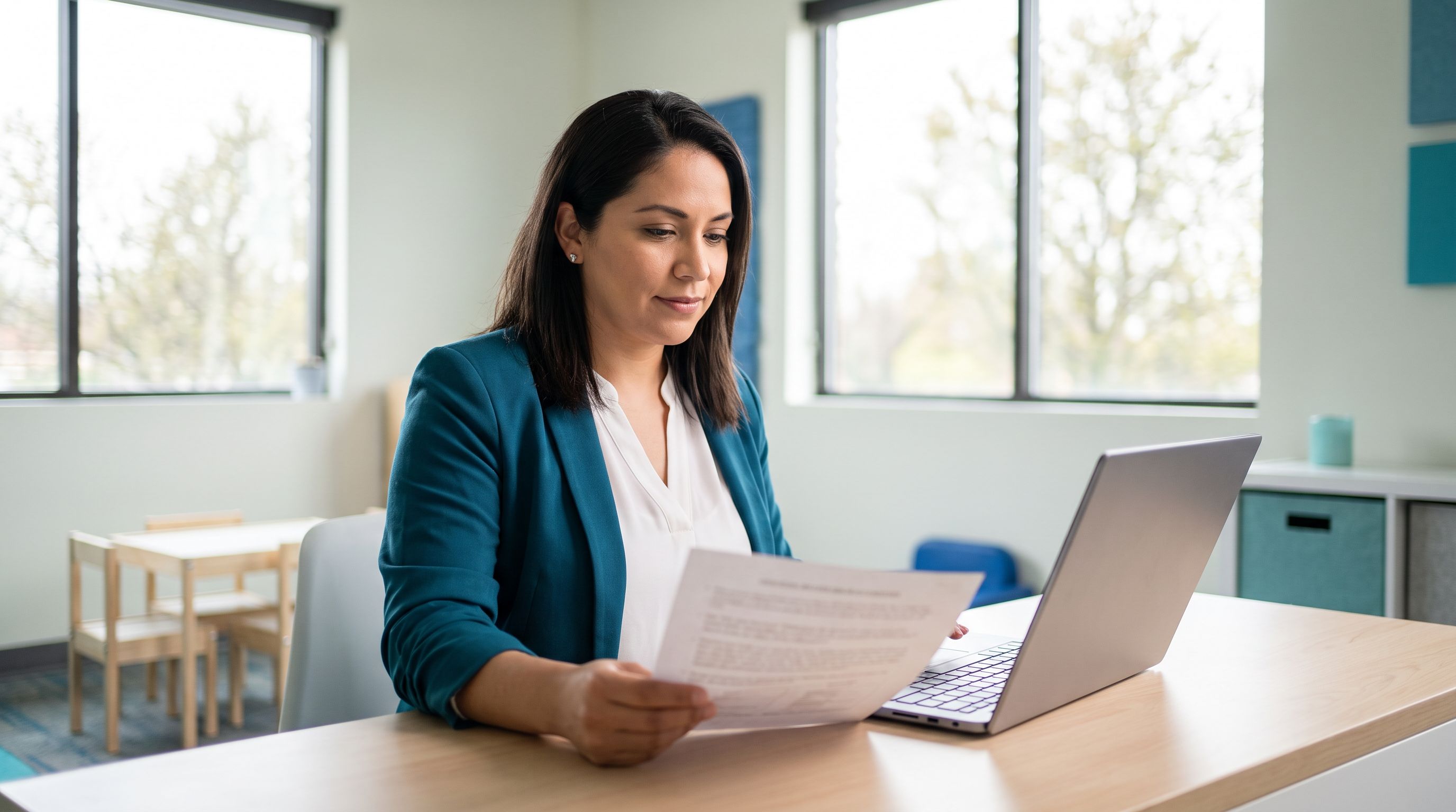 A Latina board certified behavior analyst in a teal blazer reviews a printed job offer letter beside an open laptop in a bright suburban Chicago ABA clinic.