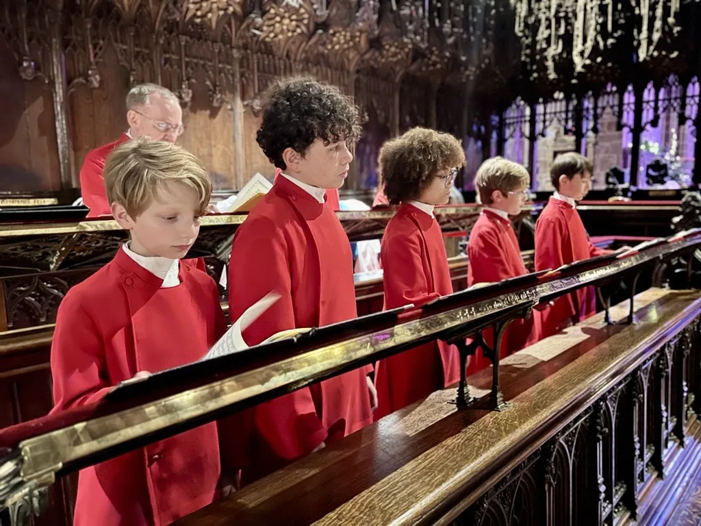 Boy choristers and Lay Clerks - both in red cassocks - stand in the Quire Stalls of Chester Cathedral. Facing the Director of Music (away from camera) some look down at their music and some look at the Director.