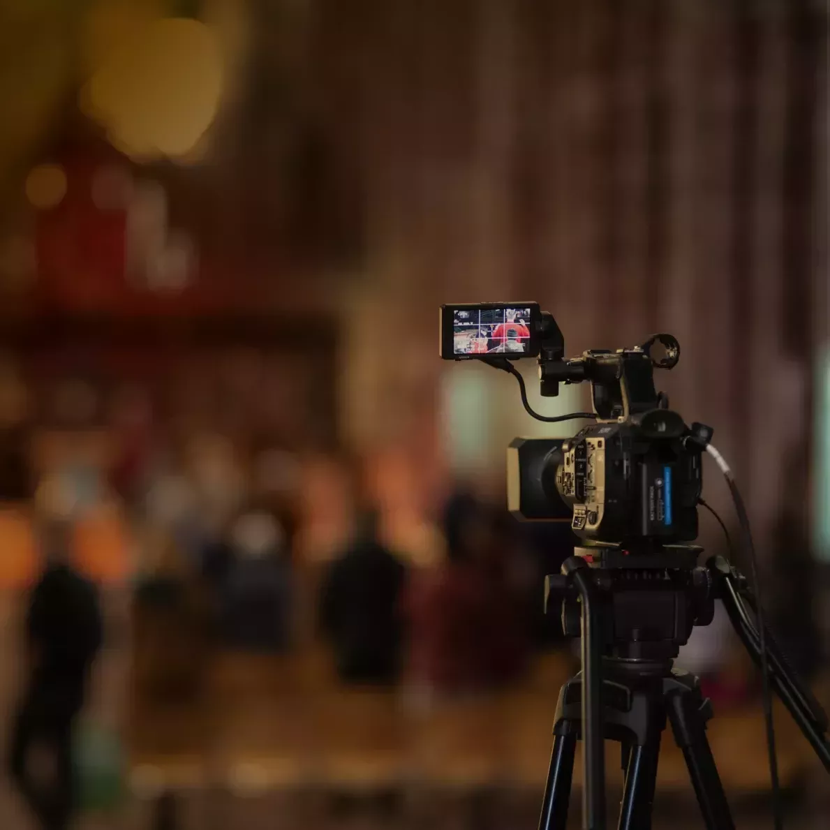 Photograph showing a video camera filming a service. The photo is taken with short depth of field with the camera in focus and the rest of the scene out of focus.