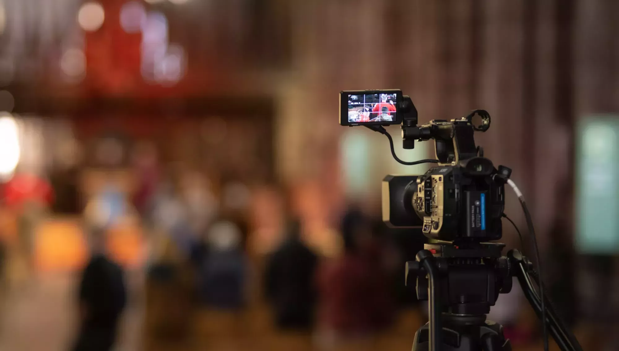 Photograph showing a video camera filming a service. The photo is taken with short depth of field with the camera in focus and the rest of the scene out of focus.