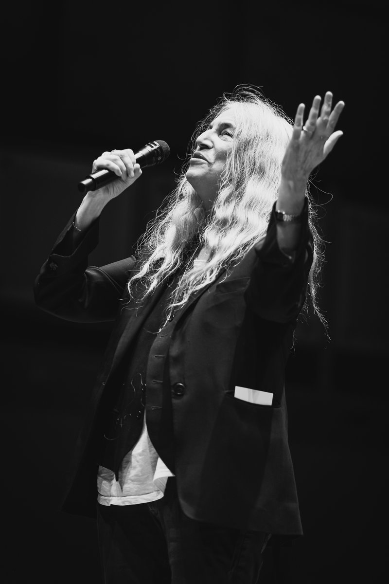 Alt-text: A black and white photo of Patti Smith singing into a microphone, with head and arms raised. She has long white hair and wears a baggy black suit.]