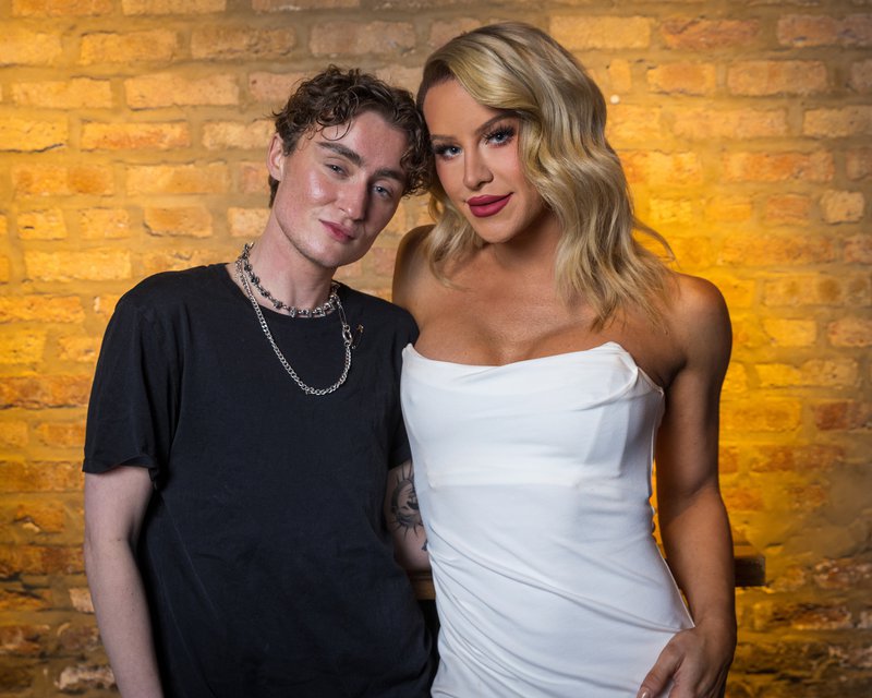 Transgender man Gottmik in a black tee and silver chains and transgender woman Gigi Gorgeous in a drapey white tube top and glamorous makeup pose arm in arm against a brick wall with a glowing yellow light behind them.