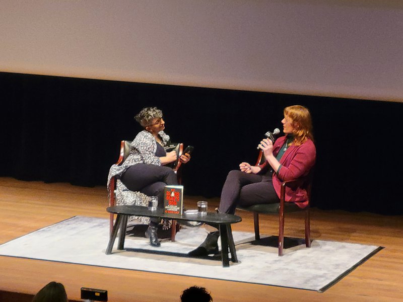Two women sit on stage, leaned in engagedly to each other. The woman to the left has cropped gray curly hair and a long blue and white cardigan. The woman to the right speaks into her mic; she has shoulder length blonde hair and a red blazer of black leggings.