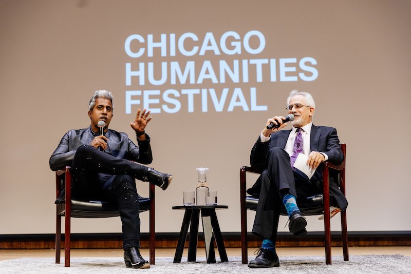 Authors Giridiharadas and Corn sit cross legged in chairs, holding mics and smiling at audience. White text projected at back reads Chicago Humanities Festival. Giridiharadas wears a black leather jacket; Corn wears a dark suit and bright socks.