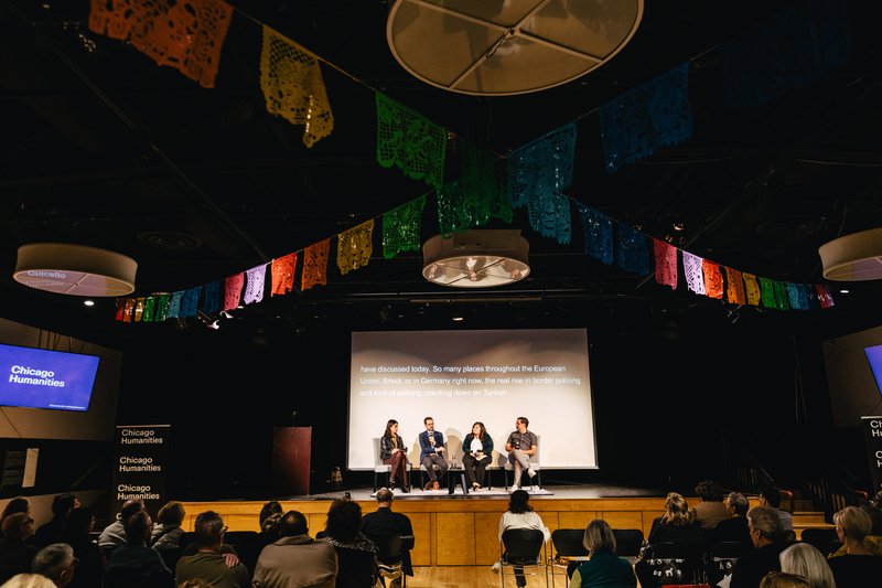 A photo from the back of a small theater, with rows of people sitting in chairs looking at four speakers up on stage. Colorful papel picado bunting criss-crosses across the ceiling.
