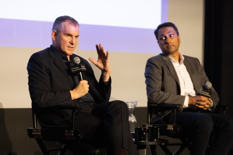 Two men sit next to each other on stage, holding microphones. To the left, a white man in all black speaks seriously and gestures with a hand. To the right, an Indian man in a gray blazer and white collared shirt looks on and listens.