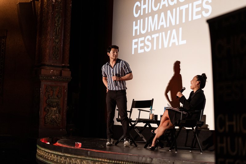 Actor Simu Liu strikes a pose, cracking up author Joanne Molinaro on an ornate movie theater stage. Holding mics, Liu wears a sharp striped tee and Molinaro wears a black suit dress. White text projected behind them reads Chicago Humanities Festival.