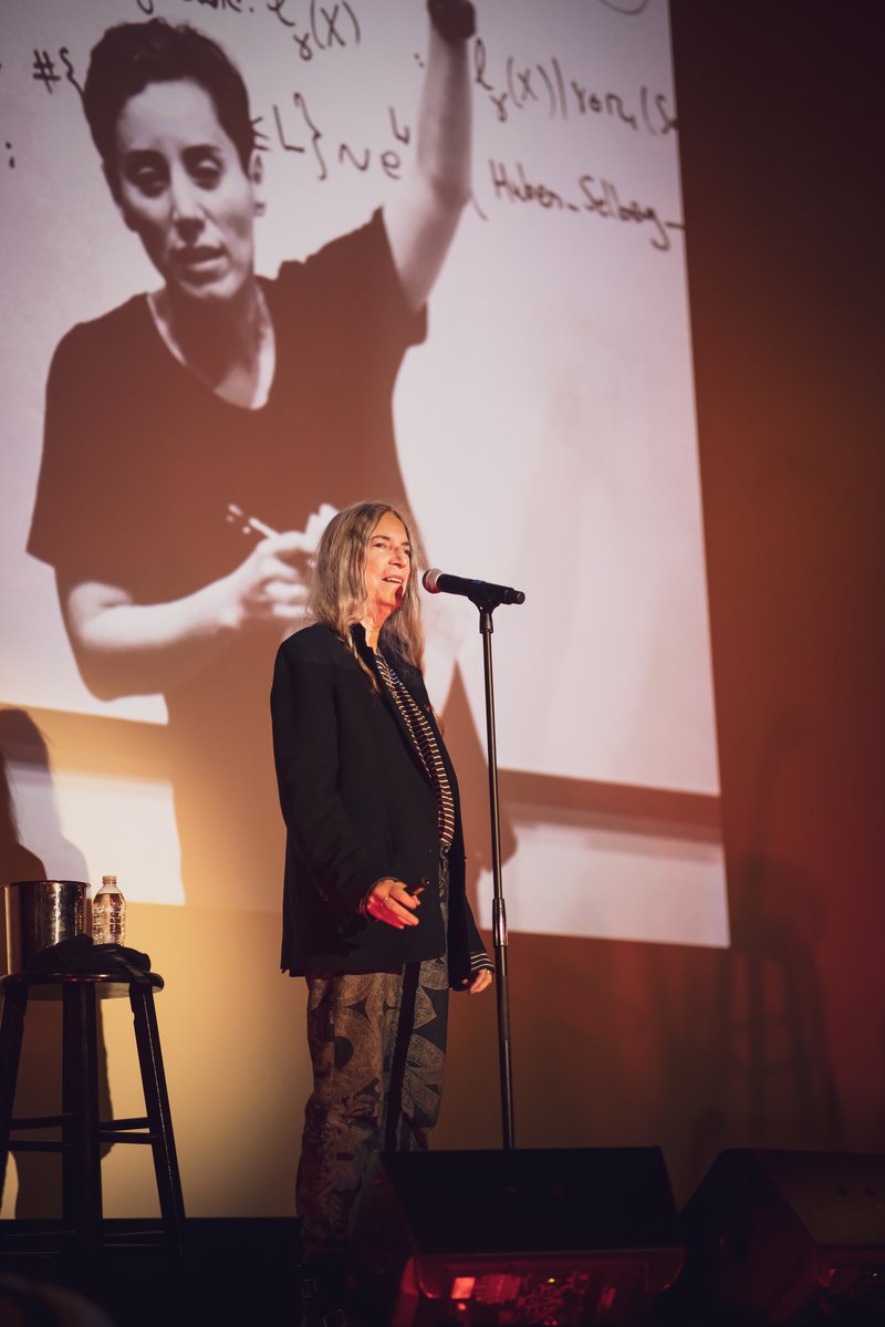 Alt-text: Patti Smith on stage at the Music Box Theatre, speaking into a microphone. She stands in front of the movie screen with a projected photo of Maryam Mirzakhani - a woman with short hair and a baggy t-shirt holding notes lecturing in front of a large math equation.