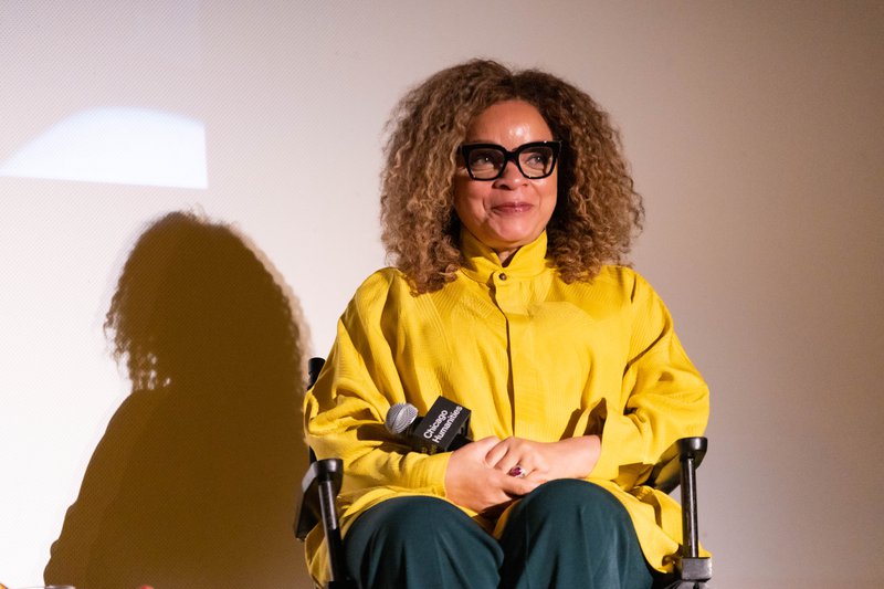 A photo of a Black woman smiling at the audience, holding a mic. She has large curly hair with bleached highlights, big black square glasses, and wears chic oversized yellow jacket and green pants.
