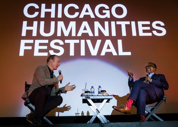 Actors Bob Odenkirk and Tim Meadows share a laugh while holding microphones and sitting in director’s chairs onstage. White text projected behind them reads Chicago Humanities Festival. Odenkirk wears a brown suede blazer while Meadows wears a blue suit and bright red socks.