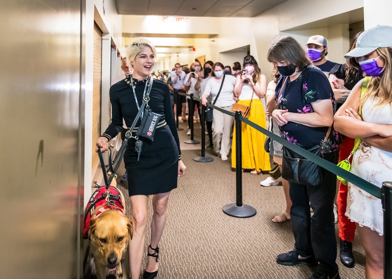 Alt-text: Selma Blair fiercely walks a hallway holding her big blond dog’s leash, saying hi to a line of adoring masked fans.]