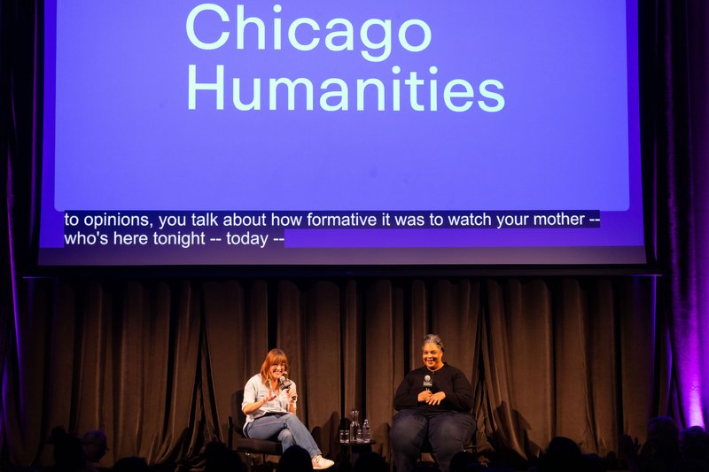 Two women sit on stage in front of a large purple screen that reads “Chicago Humanities” with live captioning underneath. They both smile at the crowd and hold microphones.