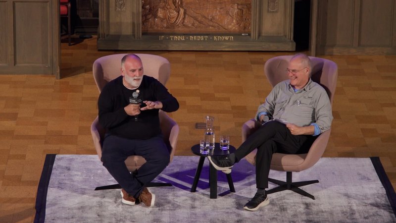 Two men holding microphones sit across from each other in funky large mauve armchairs. José Andrés sits on the left, wearing a black sweater and with a cropped white beard, speaking and gesturing. Mark Bittman to the right wears wire glasses and a casual button down, legs crossed and laughing.
