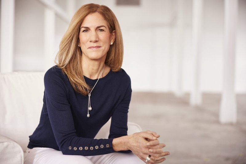 A portrait of a white woman with shoulder-length hair, a navy shirt, and a necklace with two pearls dangling from the middle. She looks warmly and confidently to the camera with her hands gently clasped on a white chair in a white room.