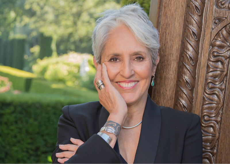 [Alt-text: Joan Baez, with cropped lush gray hair and a warm smile directly into camera, poses against an ornate wood door frame with her hand lovingly on her cheek.]