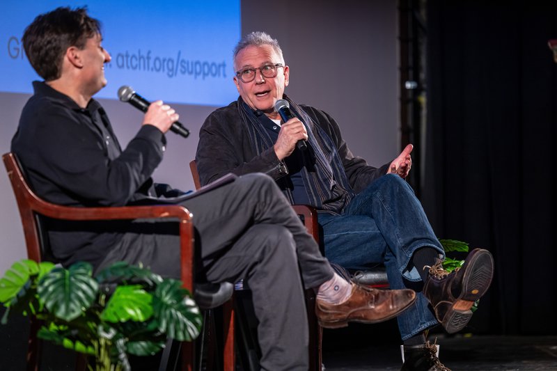 Photo of two white men on stage holding microphones. Focus is Paul Reiser to the right, with cropped gray hair and hip glasses. He wears a navy suede jacket, blue jeans, and rugged brown boots. To the left, Mark Bazer throws his head back and laughs. He wears a dark gray button down, light gray trousers, and worn brown shoes.
