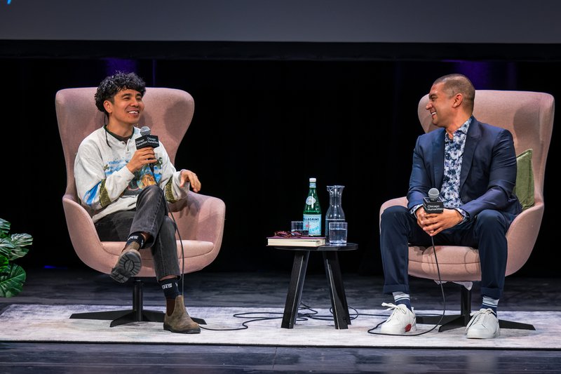 A photo of two men laughing on stage. The Vietnamese American man to the left is mid-sentence, holding a microphone. He has shaggy black bowl cut hair with a long rattail braid behind one ear. He wears a retro white sweatshirt, black jeans, and Blundstone boots. The man to the right has a buzzed head, and wears a sharp blue suit, a white and blue floral shirt, and white sneakers.