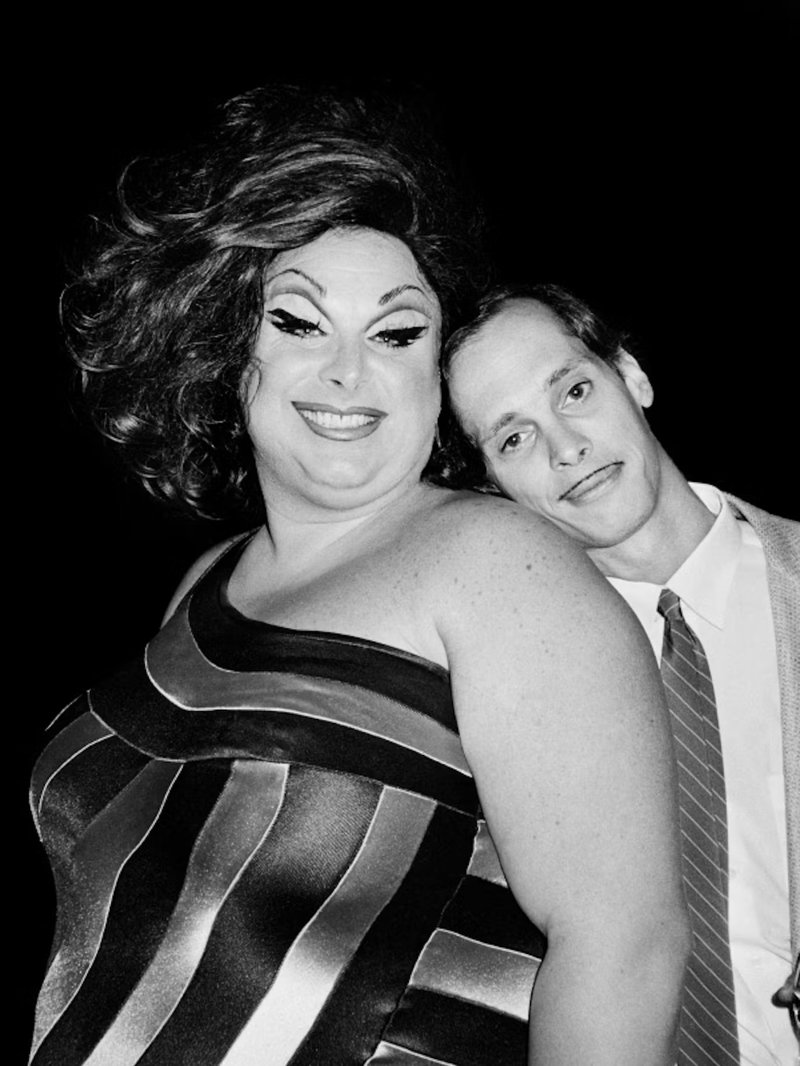 Black and white photo of Divine in a big ‘80s wig parted to one side and a shiny striped dress with John Waters resting his head on her shoulder.