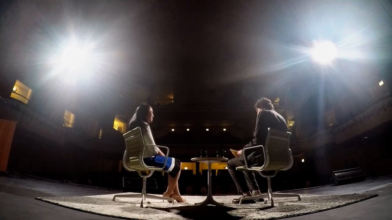 Alt text: A photo taken from the back of the stage – a woman to the left and a man to the right sit in white chairs in front of the audience, with bright lights shining on them.
