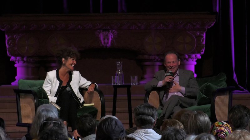A photo of Gloria Groom and Adam Gopnik sharing a laugh. They sit in green velvet chairs on stage. Behind them, an ornate stone wall carving with a neon purple light shining on it.