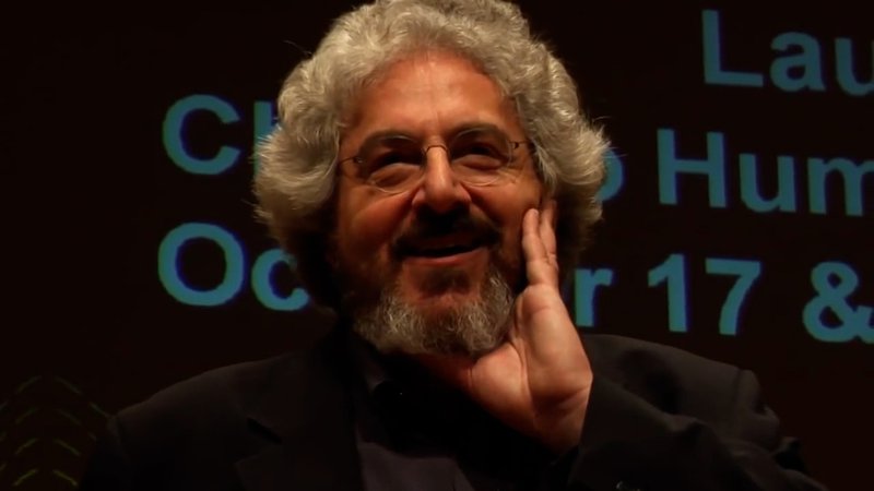 Alt-text: Screengrab of Harold Ramis, fluffy white hair and beard with wire-rimmed glasses, smiling and touching his face with a happy memory.
