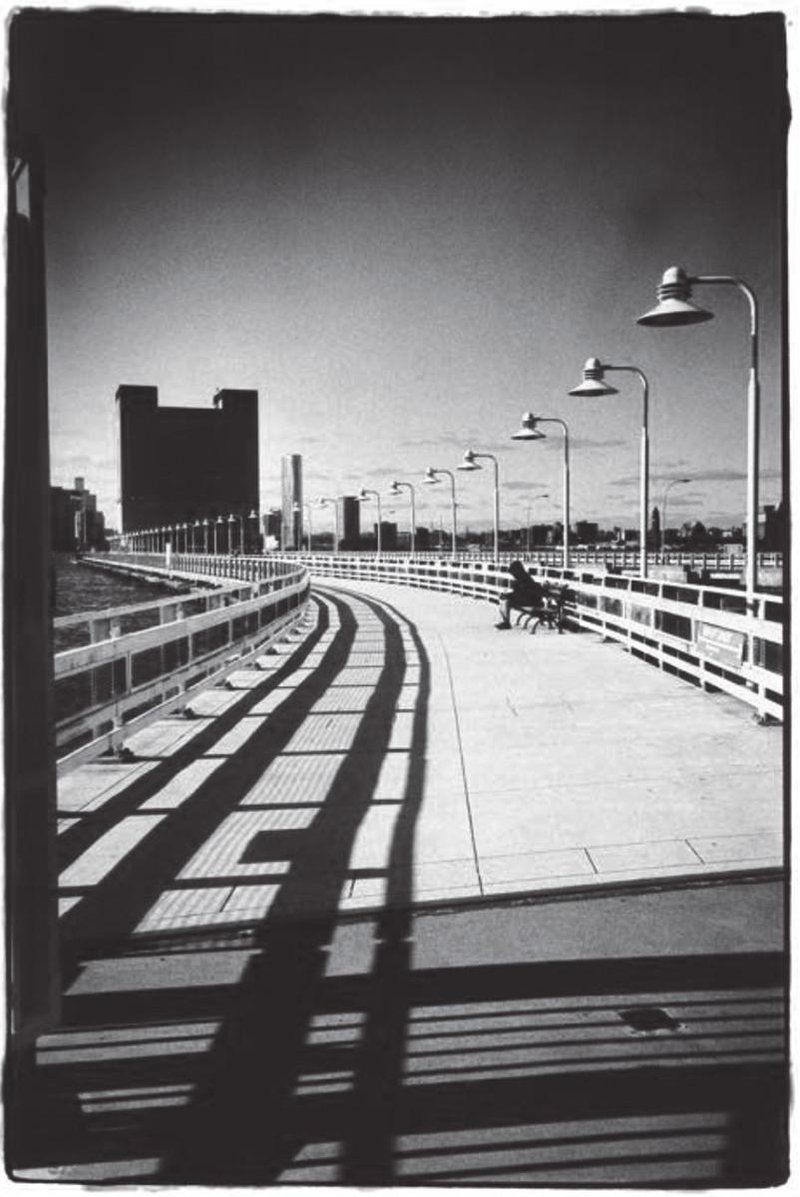 alt text: A black and white photo of a pier. The railing shadow winds around one side, lamps border the other. A person sits on a bench.