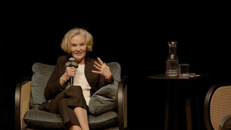 Jessica Lange elegantly sits in a plush green chair, holding a microphone and gesturing, looking to audience with a smile.
