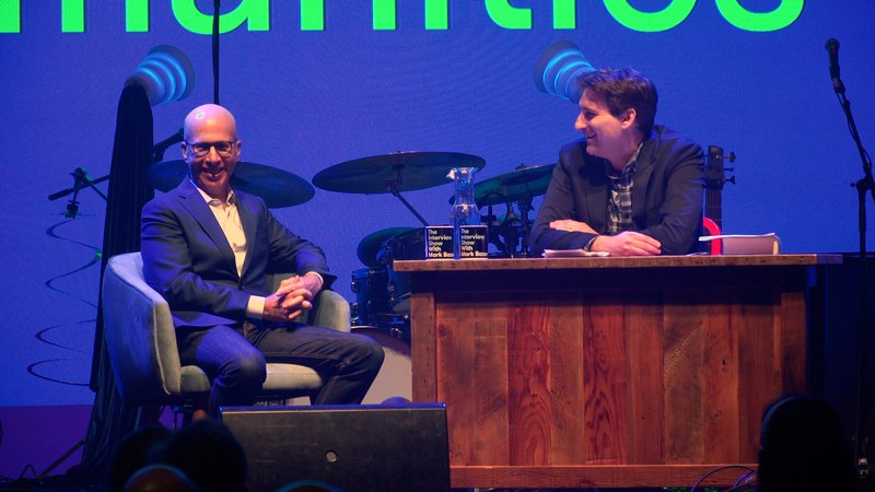 A man with glasses sits in a cushy gray chair on stage, in front of a drum kit. He wears a nice blue suit and smiles toward the audience with his hands clasped. His interviewer has shaggy hair and sits behind a desk, smiling at him with his arms folded.
