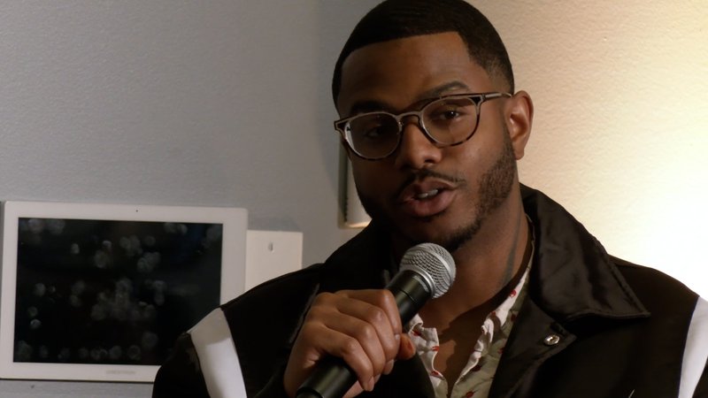 Closeup on a Black man talking confidently into a mic. He wears tortoise-rimmed glasses and has a short manicured beard.