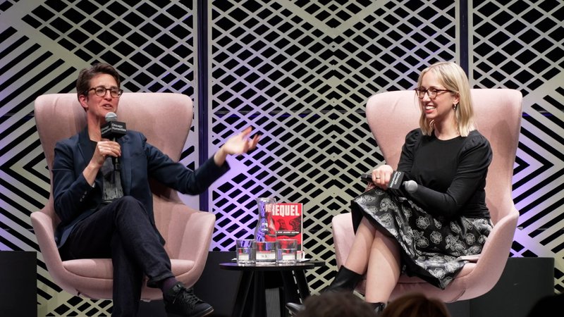 Alt text: Two women sit on stage in funky large light pink chairs. Rachel Maddow, to the left, has short hair and glasses, and wears a dark suit. She smiles and gestures. Kathleen Belew, right, has shoulder-length brown hair, glasses, and wears a black and white patterned skirt. She smiles.