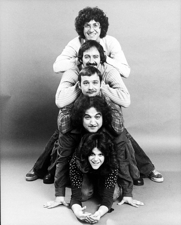 Alt-text: Black and white photo of the five young actors stacked on each others’ backs, dressed in ‘70s clothes.