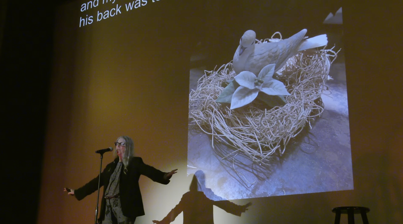 Alt-text: Patti Smith on stage at the Music Box Theatre, speaking into a microphone. She stands in front of the movie screen with a projected photo of a porcelain bird and flower in a realistic nest.