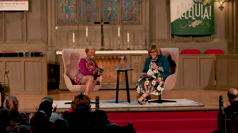 Two Black women sit in abstract pink chairs on stage at a large church. The woman to the left smiles at the audience, with cropped hair and a long sleeve fuschia  floral mini dress. The woman to the right speaks into a microphone; she has a blond bob, black glasses, and wears a denim jacket over a long floral dress. Behind them, a crucifix, candles, and a tapestry that reads “Alleluia!”