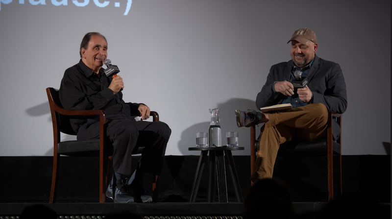 Two white men on stage, smiling into handheld microphones and sitting in large brown wooden chairs. Stine to the left wears all black, Borrelli to the right wears a navy sportcoat and khaki pants.