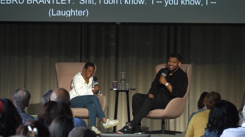 Two people share a laugh on stage, sitting in large pink chairs and holding microphones in front of an audience. To the left, a Black woman with cropped hair, delicate gold jewelry, a flowy top, and jeans. To the right, a Black man with a cropped beard, wearing all black.