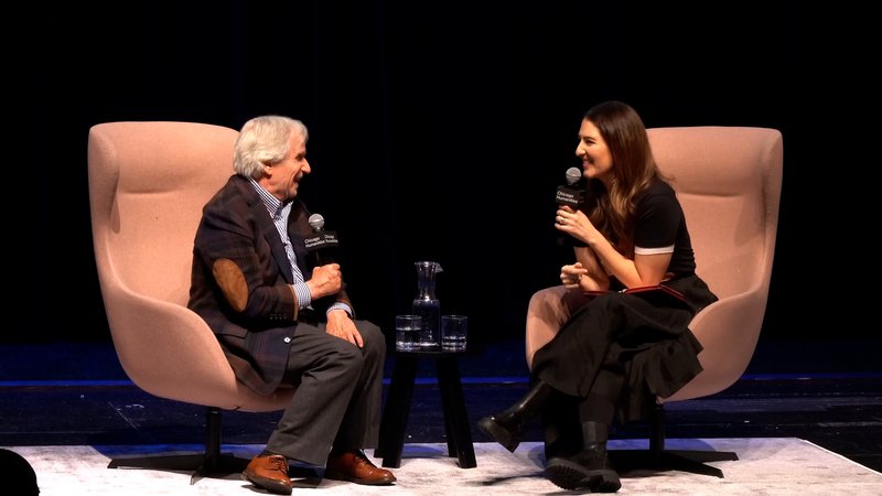 Two people on stage holding microphones, facing and laughing at each other. A man, left, with gray hair, plaid blazer, and shiny brown shoes beams at a woman, right with long brown hair and a long black dress.