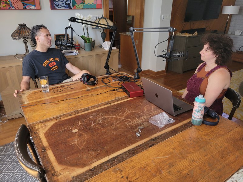 A latino man and a white woman sit at a rustic table with engravings, in the middle of an artistic house. They speak into professional microphones clamped to the table. The man has cropped dark curly hair and wears a t-shirt, the woman has curly brown hair and wears purple overalls. / Un hombre latino y una mujer blanca se sientan en una mesa rústica con grabados en medio de una casa artística. Hablan con micrófonos profesionales sujetos a la mesa. El hombre tiene el pelo oscuro y rizado y viste una camiseta.  La mujer tiene el pelo castaño rizado y viste un overol morado.